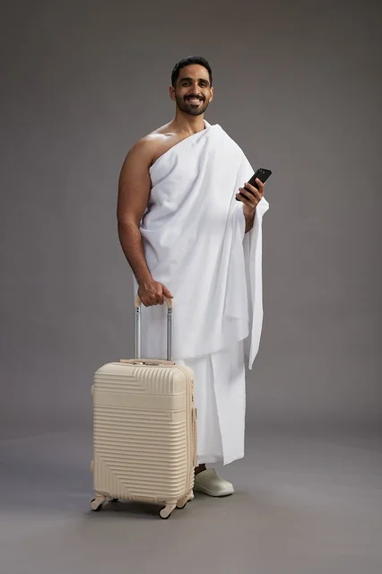 A portrait of an Arab Saudi Gulf man in Ihram clothing with a travel bag, using a mobile phone, going to Mecca for Hajj and Umrah, full body, gray background.
