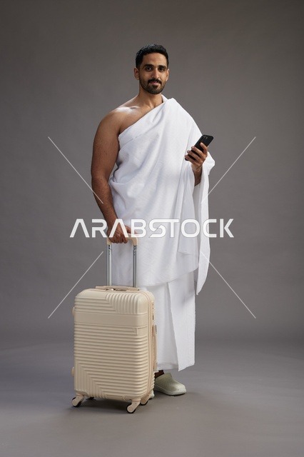 بورتريه لرجل عربي  سعودي خليجي بملابس الإحرام مع حقيبة سفر, استخدام الهاتف المحمول, الذهاب الى مكه للحج والعمره, كامل الجسم, خلفيه رماديه