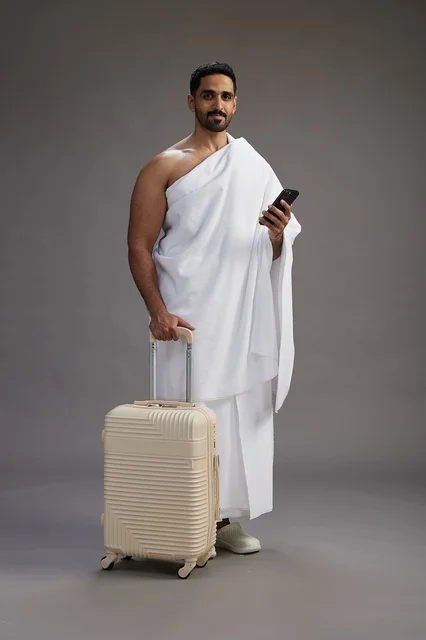 A portrait of an Arab Saudi Gulf man in Ihram clothing with a travel bag, using a mobile phone, going to Mecca for Hajj and Umrah, full body, gray background.