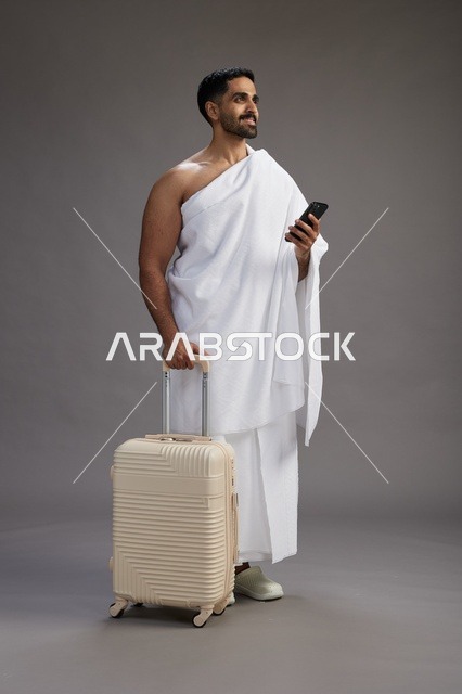 بورتريه لرجل عربي  سعودي خليجي بملابس الإحرام مع حقيبة سفر, استخدام الهاتف المحمول, الذهاب الى مكه للحج والعمره, كامل الجسم, خلفيه رماديه