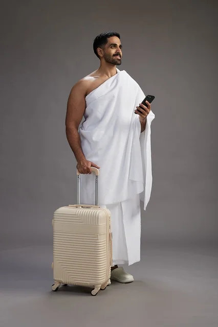 A portrait of an Arab Saudi Gulf man in Ihram clothing with a travel bag, using a mobile phone, going to Mecca for Hajj and Umrah, full body, gray background.