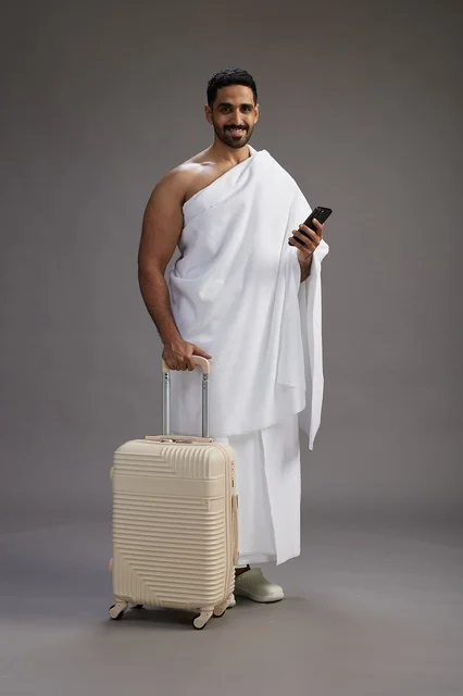 A portrait of an Arab Saudi Gulf man in Ihram clothing with a travel bag, using a mobile phone, going to Mecca for Hajj and Umrah, full body, gray background.