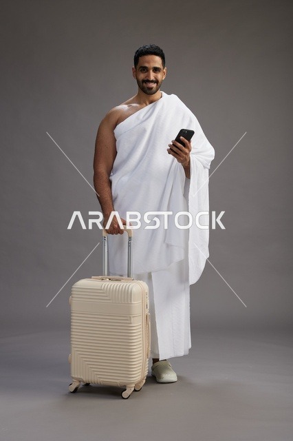 بورتريه لرجل عربي  سعودي خليجي بملابس الإحرام مع حقيبة سفر, استخدام الهاتف المحمول, الذهاب الى مكه للحج والعمره, كامل الجسم, خلفيه رماديه