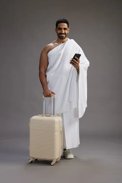 A portrait of an Arab Saudi Gulf man in Ihram clothing with a travel bag, using a mobile phone, going to Mecca for Hajj and Umrah, full body, gray background.