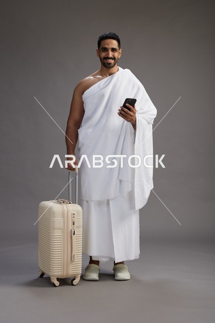 بورتريه لرجل عربي  سعودي خليجي بملابس الإحرام مع حقيبة سفر, استخدام الهاتف المحمول, الذهاب الى مكه للحج والعمره, كامل الجسم, خلفيه رماديه