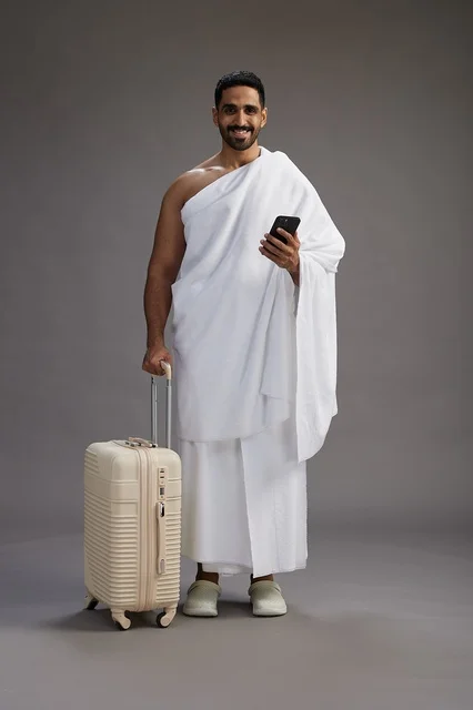 A portrait of an Arab Saudi Gulf man in Ihram clothing with a travel bag, using a mobile phone, going to Mecca for Hajj and Umrah, full body, gray background.