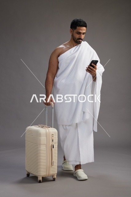 بورتريه لرجل عربي  سعودي خليجي بملابس الإحرام مع حقيبة سفر, استخدام الهاتف المحمول, الذهاب الى مكه للحج والعمره, كامل الجسم, خلفيه رماديه