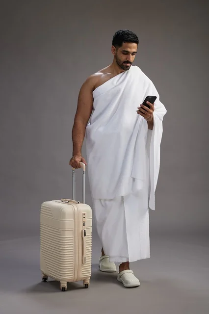 A portrait of an Arab Saudi Gulf man in Ihram clothing with a travel bag, using a mobile phone, going to Mecca for Hajj and Umrah, full body, gray background.
