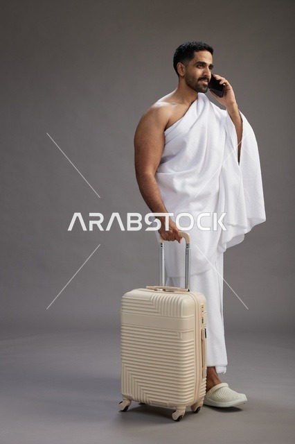 بورتريه لرجل عربي  سعودي خليجي بملابس الإحرام مع حقيبة سفر, استخدام الهاتف المحمول, الذهاب الى مكه للحج والعمره, كامل الجسم, خلفيه رماديه