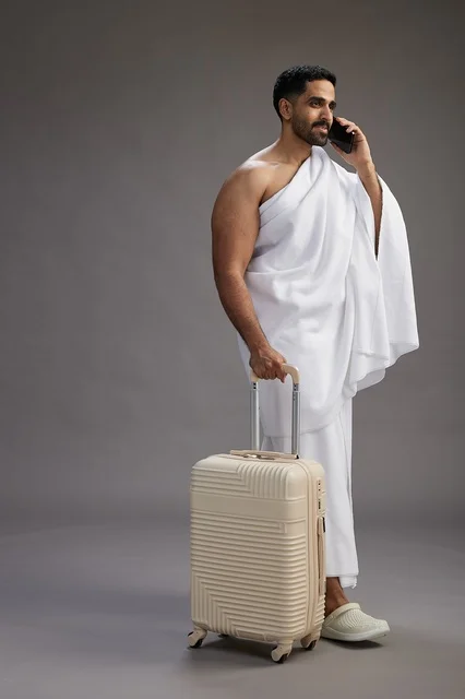 بورتريه لرجل عربي  سعودي خليجي بملابس الإحرام مع حقيبة سفر, استخدام الهاتف المحمول, الذهاب الى مكه للحج والعمره, كامل الجسم, خلفيه رماديه