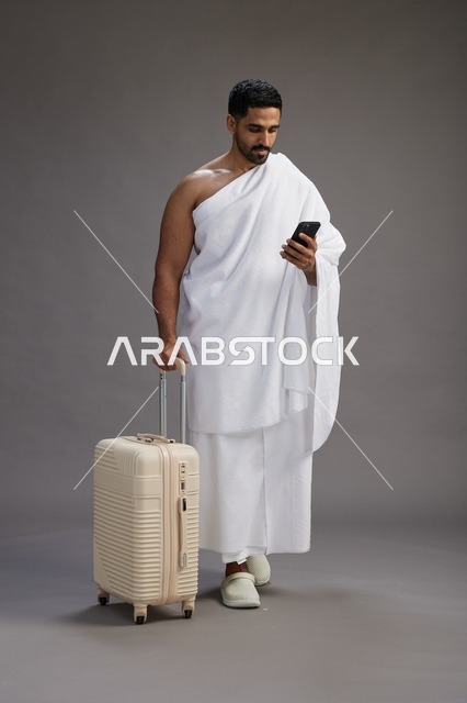 بورتريه لرجل عربي  سعودي خليجي بملابس الإحرام مع حقيبة سفر, استخدام الهاتف المحمول, الذهاب الى مكه للحج والعمره, كامل الجسم, خلفيه رماديه