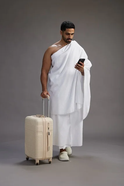 A portrait of an Arab Saudi Gulf man in Ihram clothing with a travel bag, using a mobile phone, going to Mecca for Hajj and Umrah, full body, gray background.