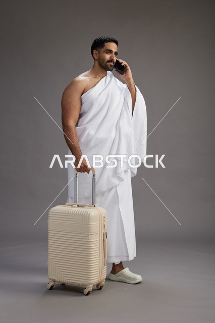 بورتريه لرجل عربي  سعودي خليجي بملابس الإحرام مع حقيبة سفر, استخدام الهاتف المحمول, الذهاب الى مكه للحج والعمره, كامل الجسم, خلفيه رماديه