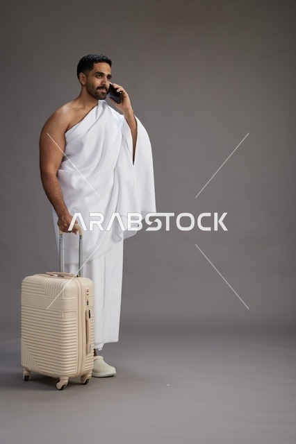 بورتريه لرجل عربي  سعودي خليجي بملابس الإحرام مع حقيبة سفر, استخدام الهاتف المحمول, الذهاب الى مكه للحج والعمره, كامل الجسم, خلفيه رماديه