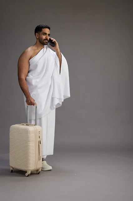 بورتريه لرجل عربي  سعودي خليجي بملابس الإحرام مع حقيبة سفر, استخدام الهاتف المحمول, الذهاب الى مكه للحج والعمره, كامل الجسم, خلفيه رماديه