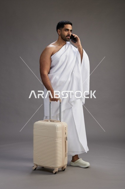 A portrait of an Arab Saudi Gulf man in Ihram clothing with a travel bag, using a mobile phone, going to Mecca for Hajj and Umrah, full body, gray background.