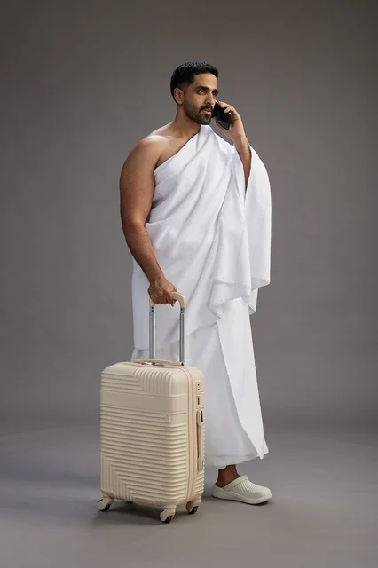 A portrait of an Arab Saudi Gulf man in Ihram clothing with a travel bag, using a mobile phone, going to Mecca for Hajj and Umrah, full body, gray background.