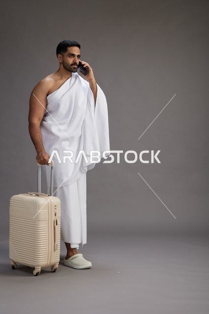 A portrait of an Arab Saudi Gulf man in Ihram clothing with a travel bag, using a mobile phone, going to Mecca for Hajj and Umrah, full body, gray background.