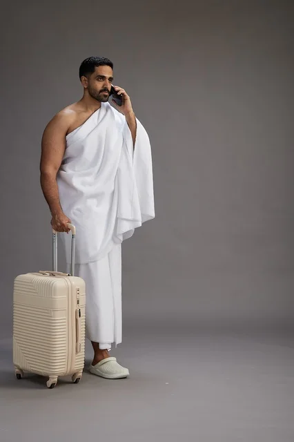 A portrait of an Arab Saudi Gulf man in Ihram clothing with a travel bag, using a mobile phone, going to Mecca for Hajj and Umrah, full body, gray background.