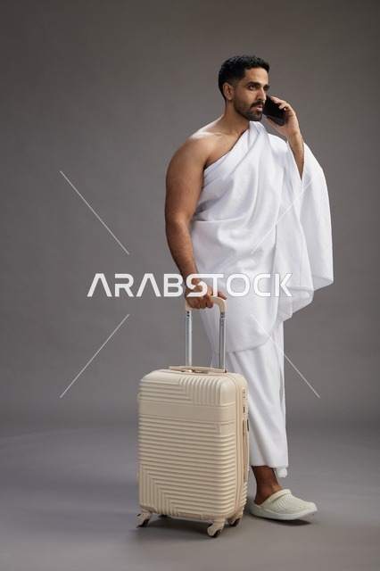 بورتريه لرجل عربي  سعودي خليجي بملابس الإحرام مع حقيبة سفر, استخدام الهاتف المحمول, الذهاب الى مكه للحج والعمره, كامل الجسم, خلفيه رماديه