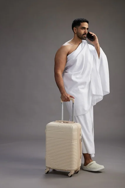 A portrait of an Arab Saudi Gulf man in Ihram clothing with a travel bag, using a mobile phone, going to Mecca for Hajj and Umrah, full body, gray background.