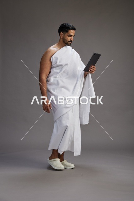 بورتريه لحاج سعودي بملابس الإحرام يحمل تابلت, الاستعداد لاداء مناسك الحج والعمره, كامل الجسم, خلفيه رماديه