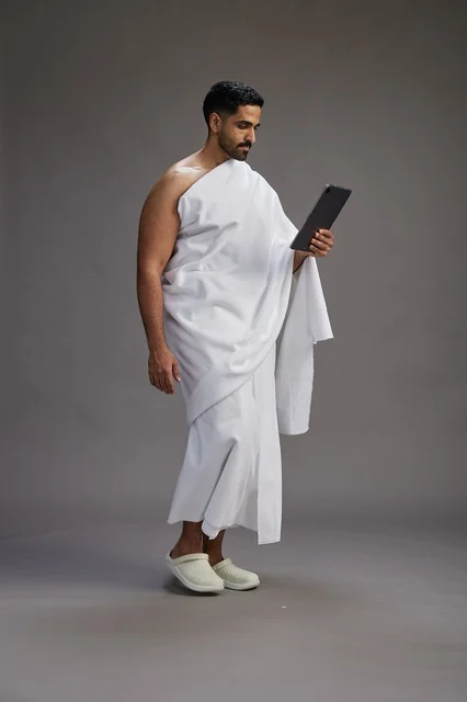 A portrait of a Saudi pilgrim in Ihram clothing holding a tablet, preparing to perform the rituals of Hajj and Umrah, full body, gray background.