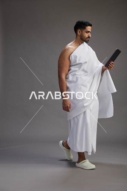 بورتريه لحاج سعودي بملابس الإحرام يحمل تابلت, الاستعداد لاداء مناسك الحج والعمره, كامل الجسم, خلفيه رماديه