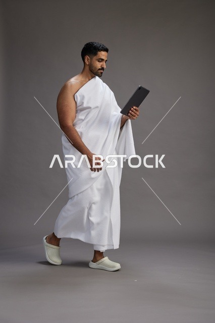 بورتريه لحاج سعودي بملابس الإحرام يحمل تابلت, الاستعداد لاداء مناسك الحج والعمره, كامل الجسم, خلفيه رماديه
