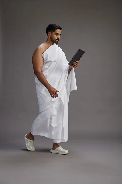 A portrait of a Saudi pilgrim in Ihram clothing holding a tablet, preparing to perform the rituals of Hajj and Umrah, full body, gray background.