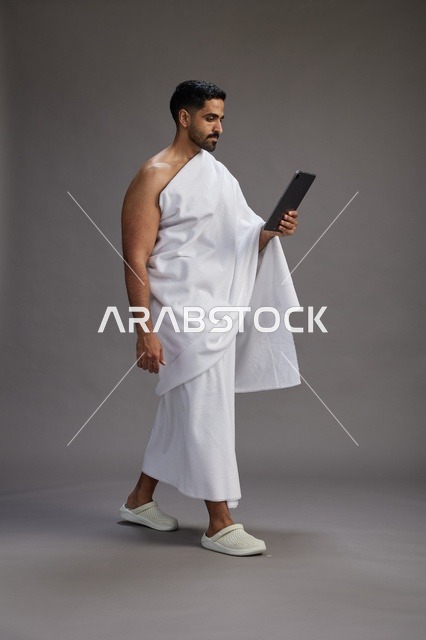 بورتريه لحاج سعودي بملابس الإحرام يحمل تابلت, الاستعداد لاداء مناسك الحج والعمره, كامل الجسم, خلفيه رماديه