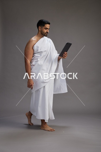 A portrait of a Saudi pilgrim in Ihram clothing holding a tablet, preparing to perform the rituals of Hajj and Umrah, full body, gray background.