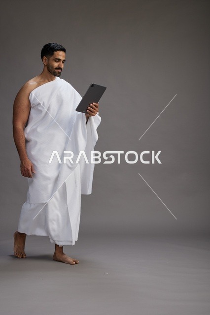 بورتريه لحاج سعودي بملابس الإحرام يحمل تابلت, الاستعداد لاداء مناسك الحج والعمره, كامل الجسم, خلفيه رماديه