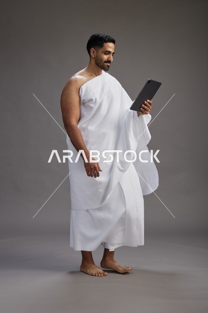 بورتريه لحاج سعودي بملابس الإحرام يحمل تابلت, الاستعداد لاداء مناسك الحج والعمره, كامل الجسم, خلفيه رماديه