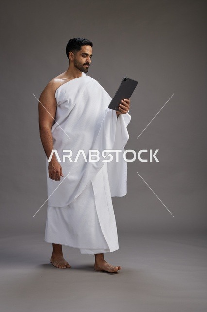 A portrait of a Saudi pilgrim in Ihram clothing holding a tablet, preparing to perform the rituals of Hajj and Umrah, full body, gray background.