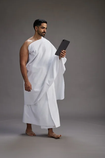 A portrait of a Saudi pilgrim in Ihram clothing holding a tablet, preparing to perform the rituals of Hajj and Umrah, full body, gray background.