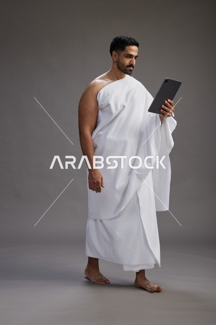 بورتريه لحاج سعودي بملابس الإحرام يحمل تابلت, الاستعداد لاداء مناسك الحج والعمره, كامل الجسم, خلفيه رماديه