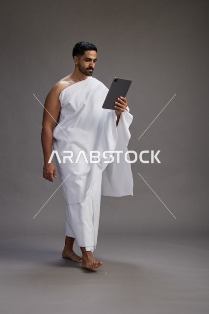A portrait of a Saudi pilgrim in Ihram clothing holding a tablet, preparing to perform the rituals of Hajj and Umrah, full body, gray background.