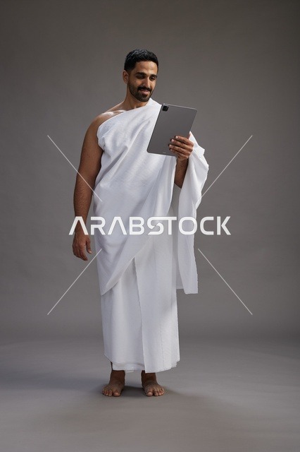 A portrait of a Saudi pilgrim in Ihram clothing holding a tablet, preparing to perform the rituals of Hajj and Umrah, full body, gray background.