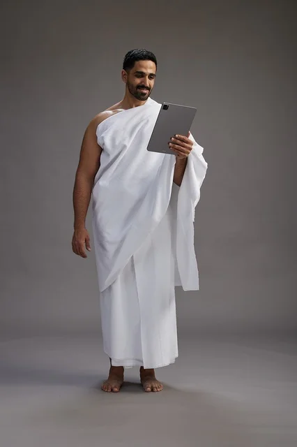 A portrait of a Saudi pilgrim in Ihram clothing holding a tablet, preparing to perform the rituals of Hajj and Umrah, full body, gray background.