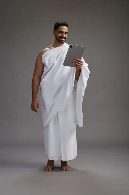 A portrait of a Saudi pilgrim in Ihram clothing holding a tablet, preparing to perform the rituals of Hajj and Umrah, full body, gray background.