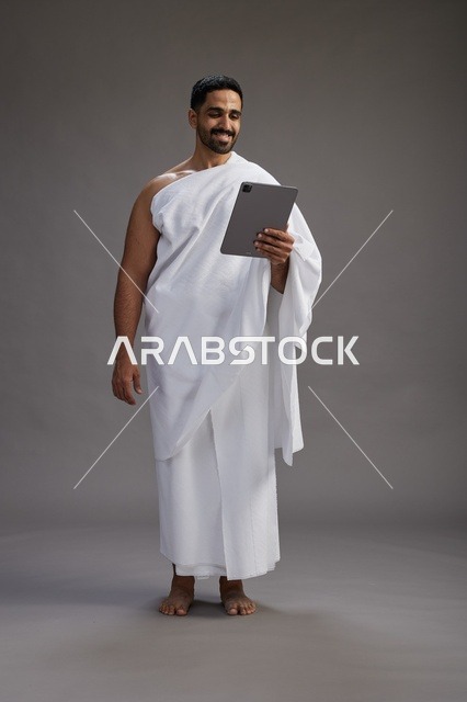 A portrait of a Saudi pilgrim in Ihram clothing holding a tablet, preparing to perform the rituals of Hajj and Umrah, full body, gray background.