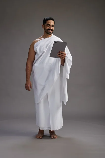 A portrait of a Saudi pilgrim in Ihram clothing holding a tablet, preparing to perform the rituals of Hajj and Umrah, full body, gray background.