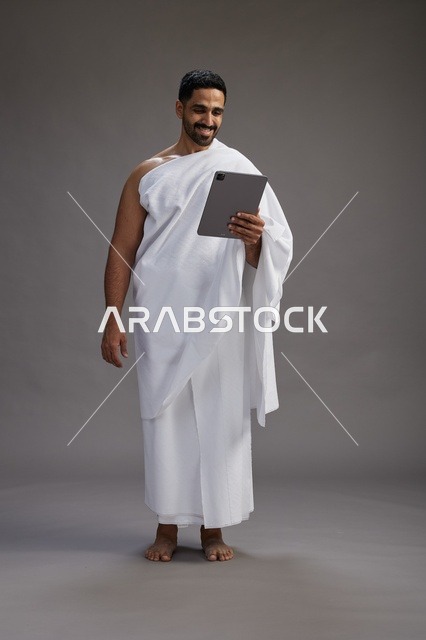 A portrait of a Saudi pilgrim in Ihram clothing holding a tablet, preparing to perform the rituals of Hajj and Umrah, full body, gray background.