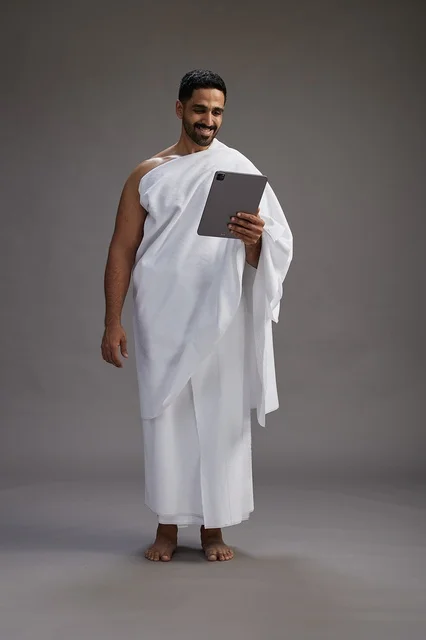 A portrait of a Saudi pilgrim in Ihram clothing holding a tablet, preparing to perform the rituals of Hajj and Umrah, full body, gray background.
