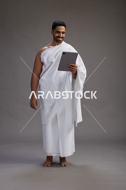 بورتريه لحاج سعودي بملابس الإحرام يحمل تابلت, الاستعداد لاداء مناسك الحج والعمره, كامل الجسم, خلفيه رماديه