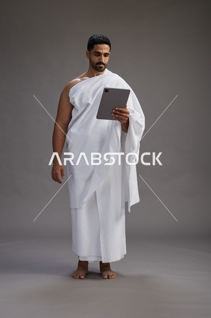 بورتريه لحاج سعودي بملابس الإحرام يحمل تابلت, الاستعداد لاداء مناسك الحج والعمره, كامل الجسم, خلفيه رماديه