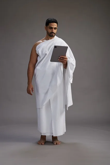 A portrait of a Saudi pilgrim in Ihram clothing holding a tablet, preparing to perform the rituals of Hajj and Umrah, full body, gray background.