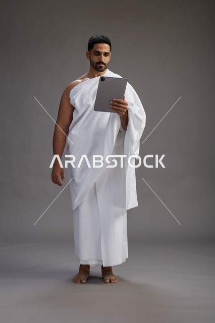 بورتريه لحاج سعودي بملابس الإحرام يحمل تابلت, الاستعداد لاداء مناسك الحج والعمره, كامل الجسم, خلفيه رماديه
