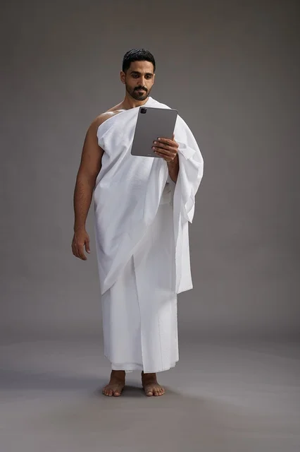 A portrait of a Saudi pilgrim in Ihram clothing holding a tablet, preparing to perform the rituals of Hajj and Umrah, full body, gray background.